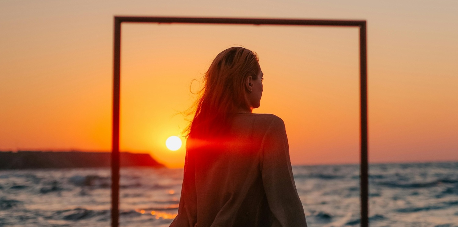 Woman sitting on beach with frame around her. Sun is setting over the ocean
